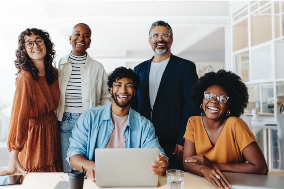 A diverse group of smiling coworkers gathered around a laptop